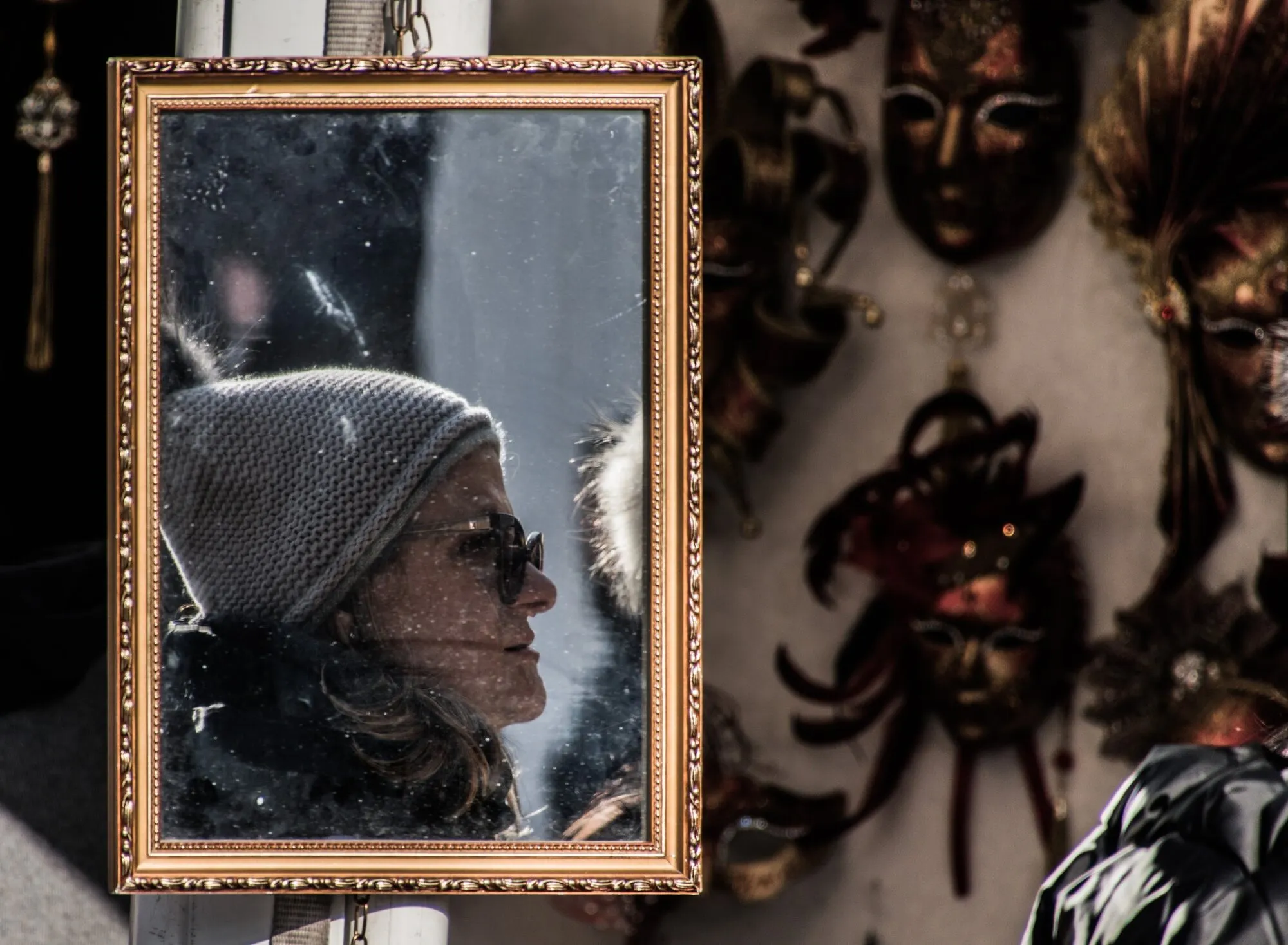 Il volto di una donna con berretto di lana e occhiali da sole riflesso in uno specchio dalla cornice dorata, accanto a una parete di maschere veneziane sfocate.