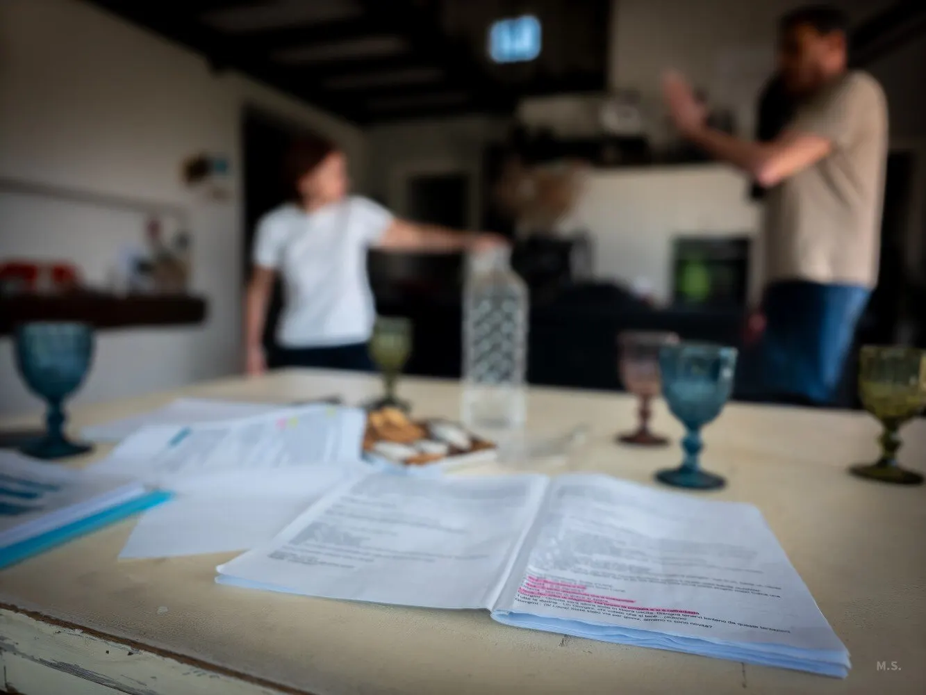 Un copione aperto con le battute evidenziate su un tavolo, tra bicchieri colorati e biscotti; sullo sfondo, fuori fuoco, due persone provano una scena.