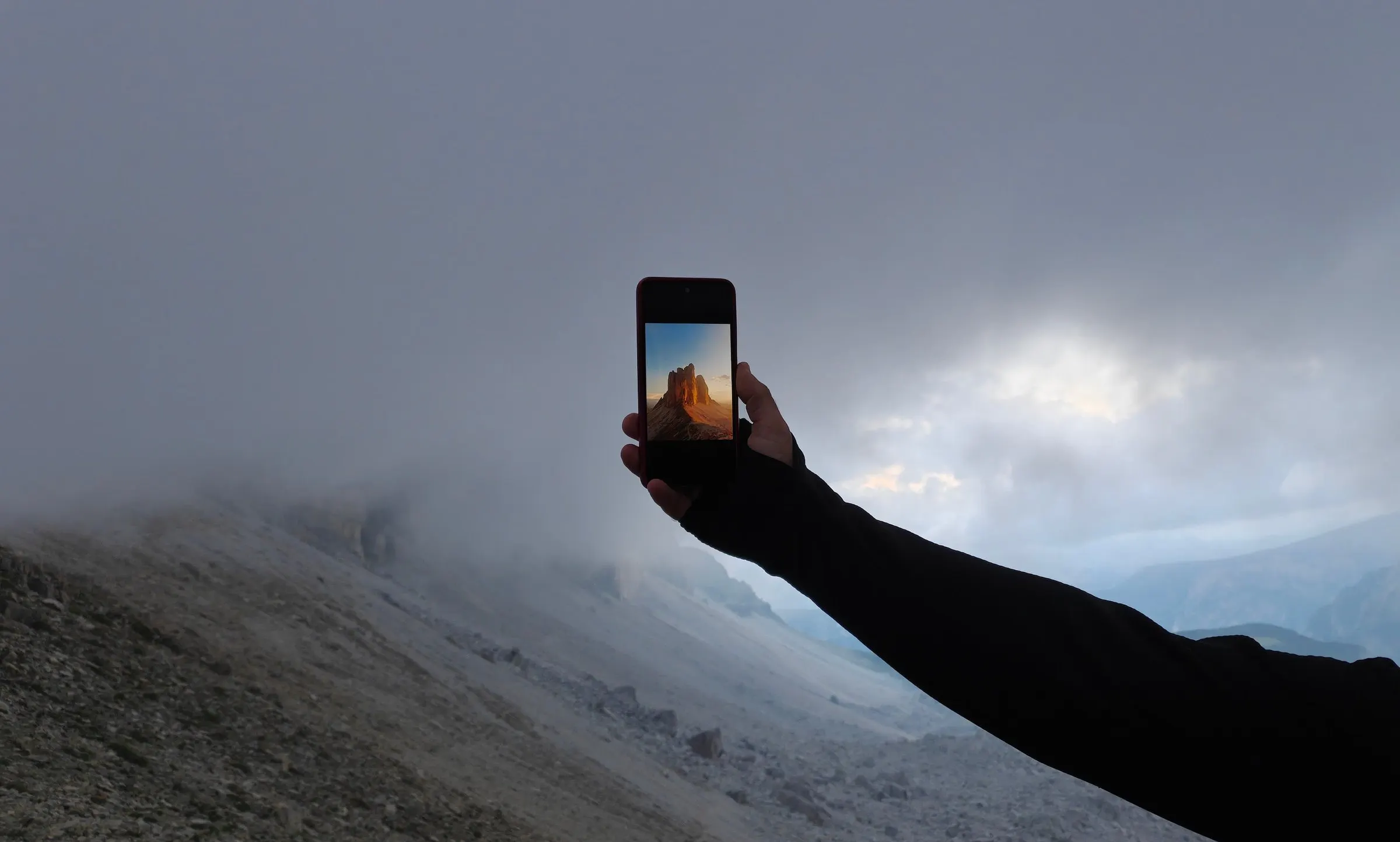 Un braccio teso nella nebbia delle Dolomiti regge uno smartphone che mostra le Tre Cime di Lavaredo illuminate dal sole