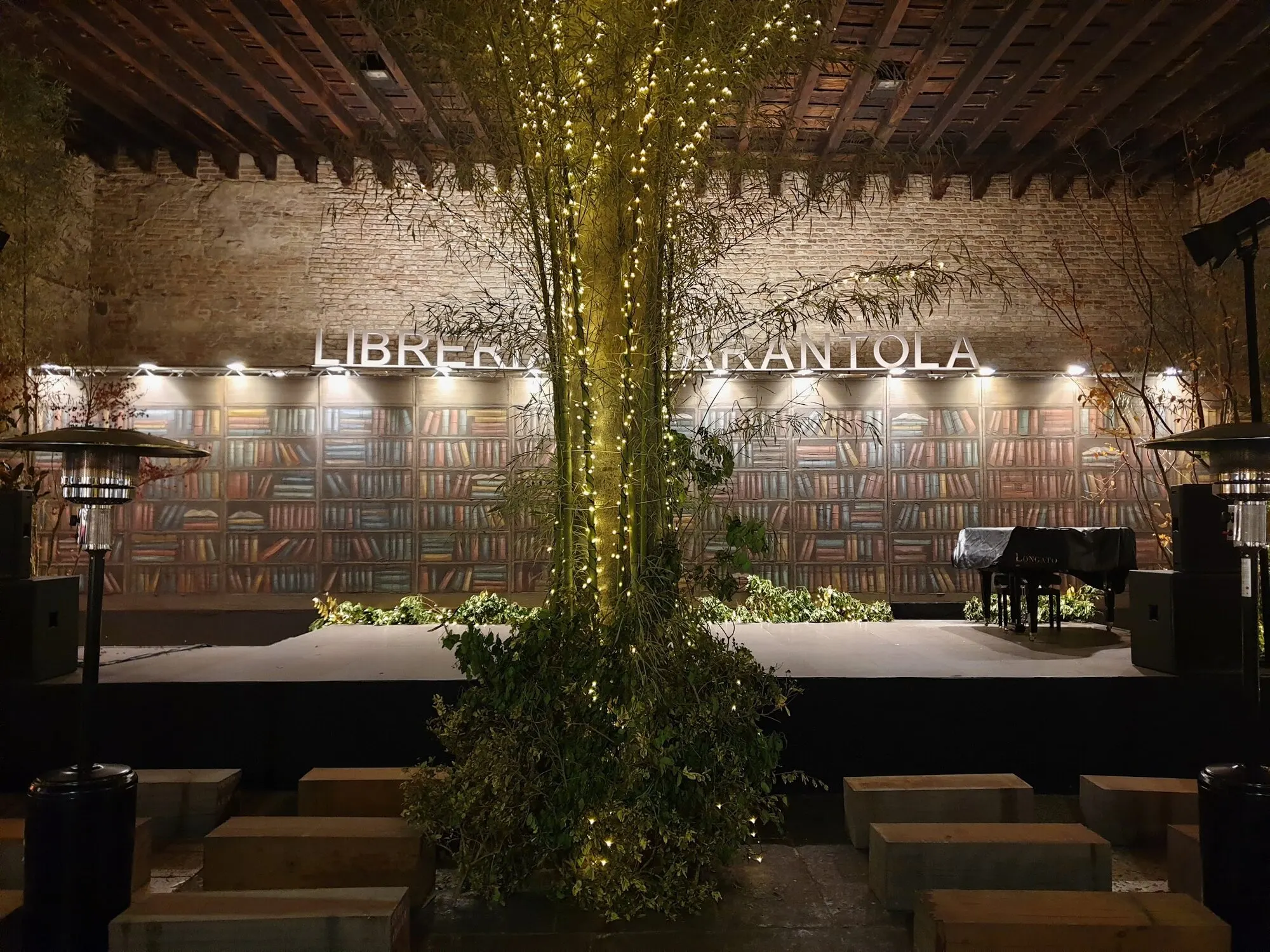 Il cortile della Libreria Tarantola di notte: una parete di mattoni con scaffali di libri dipinti, un palco con un pianoforte a coda e, al centro, un albero avvolto da piccole luci.