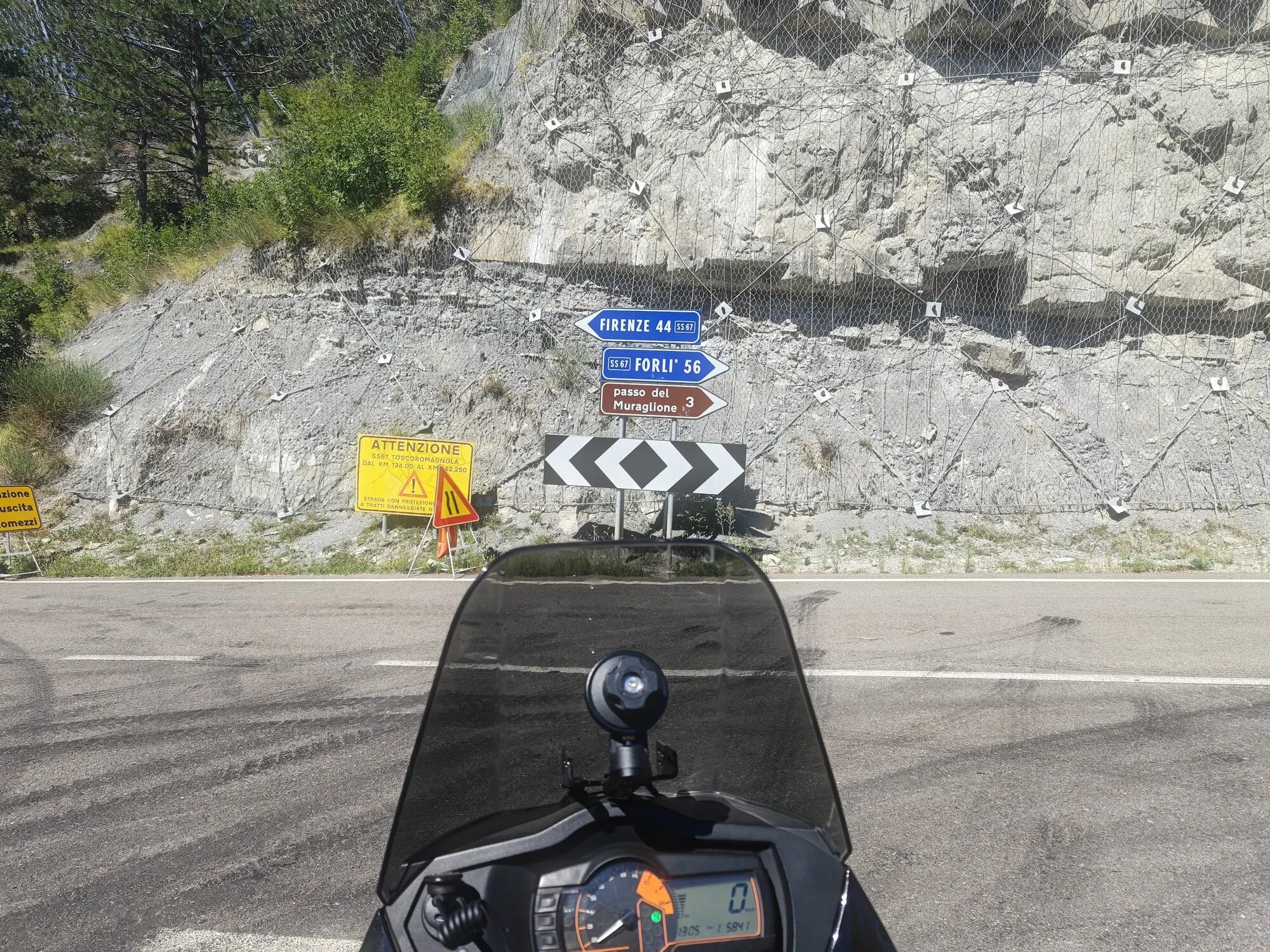 Dalla sella di una moto ferma, oltre il cupolino: un bivio sulla SS67 Tosco-Romagnola con i cartelli per Firenze (44 km), Forlì (56 km) e il Passo del Muraglione (3 km), davanti a una parete di roccia con reti paramassi.