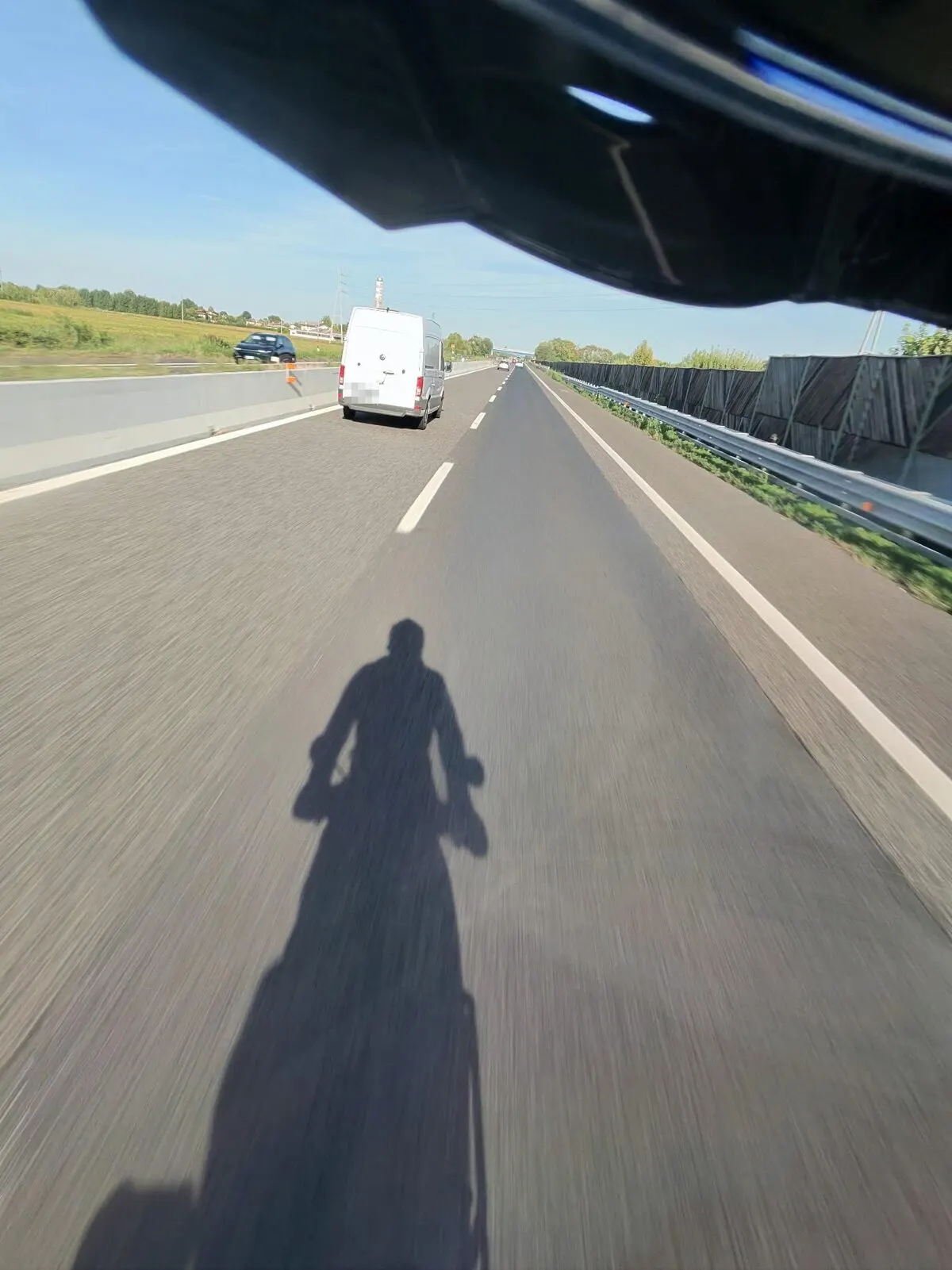 L’ombra di un motociclista in sella proiettata sull’asfalto dell’autostrada, in corsa, con un furgone bianco davanti nella corsia di sorpasso.