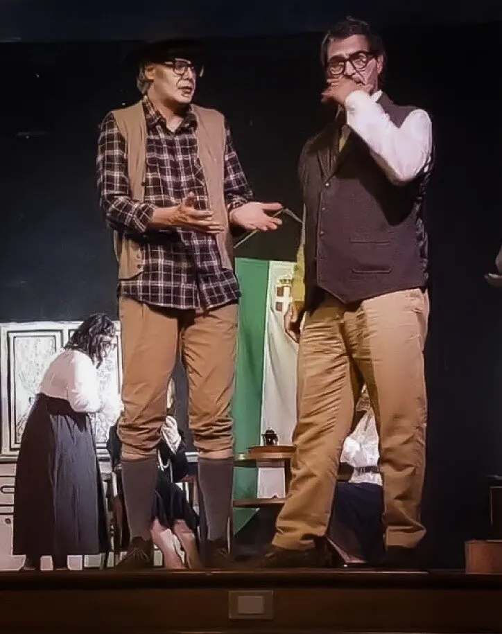 Due attori in scena con costumi d’epoca, gilet e occhiali, durante lo spettacolo; sullo sfondo una bandiera tricolore con lo stemma sabaudo e altri attori.