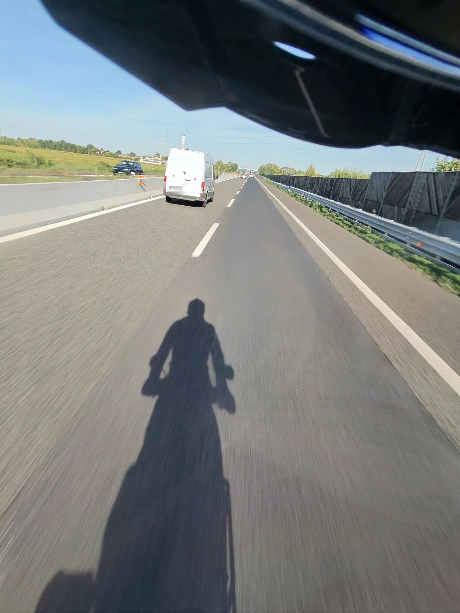 In moto su una strada a due corsie in pianura: in primo piano l’ombra del motociclista sull’asfalto, più avanti un furgone bianco.