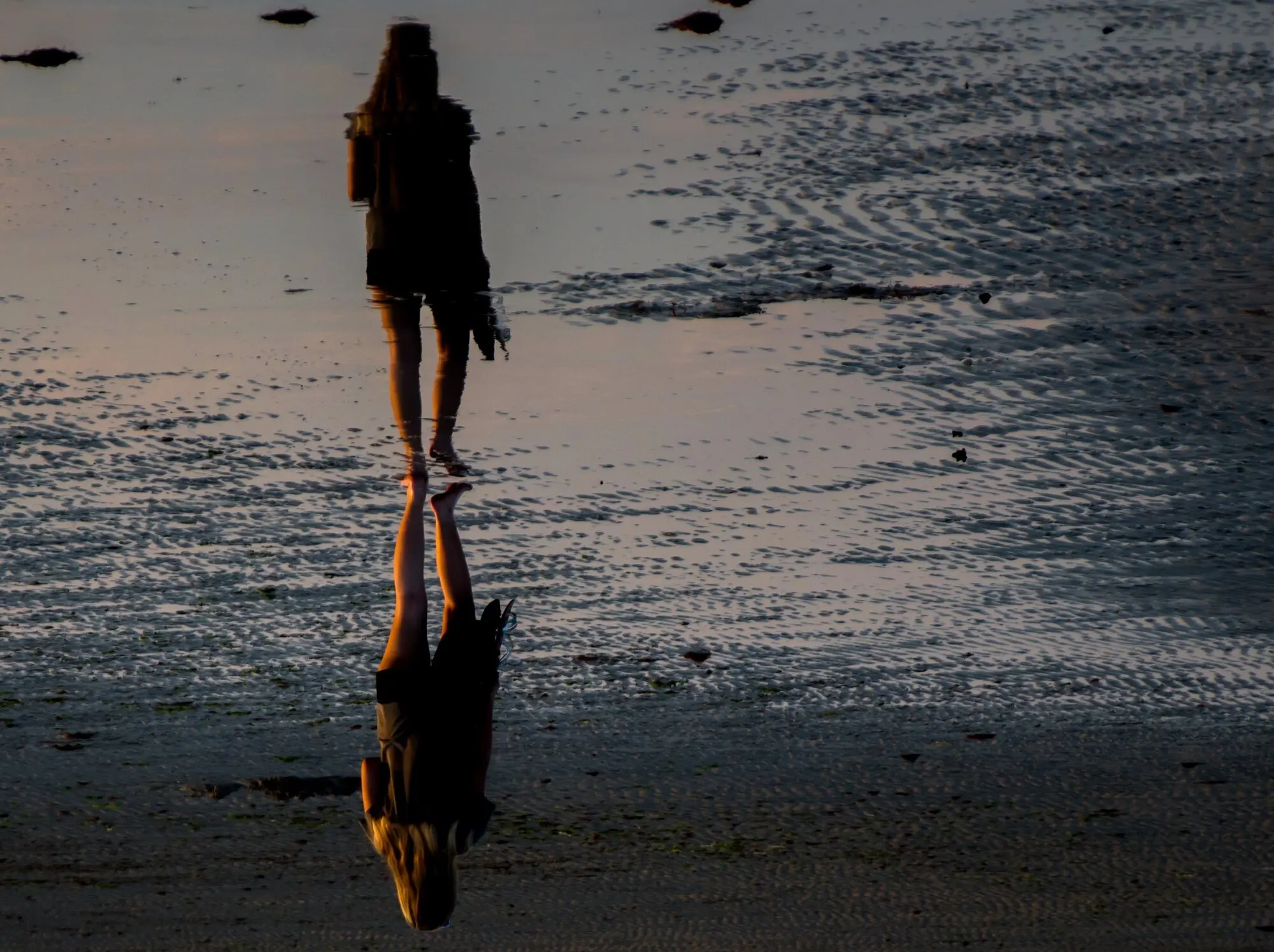 Su una spiaggia bagnata al tramonto, una persona cammina scalza e il suo riflesso capovolto si allunga sulla sabbia lucida, più nitido della figura stessa.